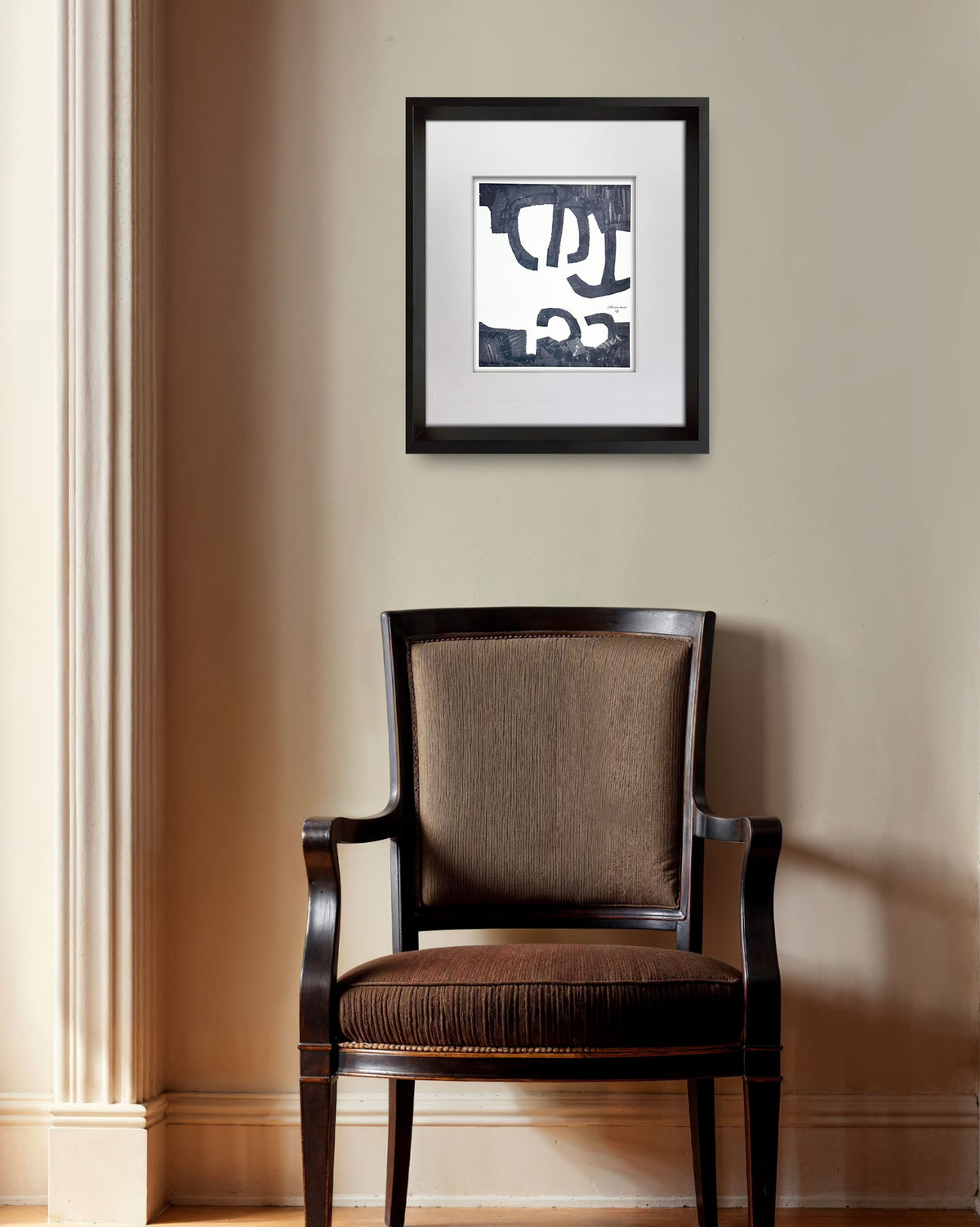A framed abstract art print with black, white, and gray shapes, displayed on a beige wall above a dark brown upholstered armchair.
