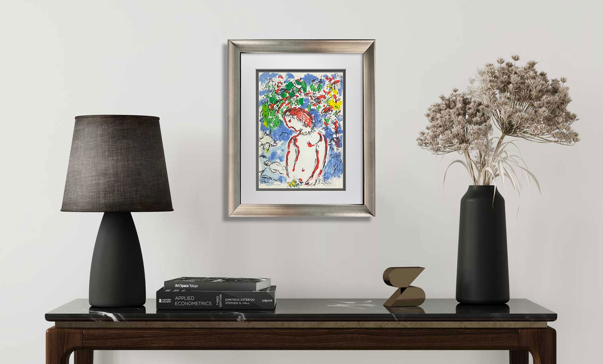 A framed colorful painting of a woman hangs on a light gray wall above a dark wooden console table styled with a black table lamp, books, a small sculptural object, and a black vase with dried flowers.