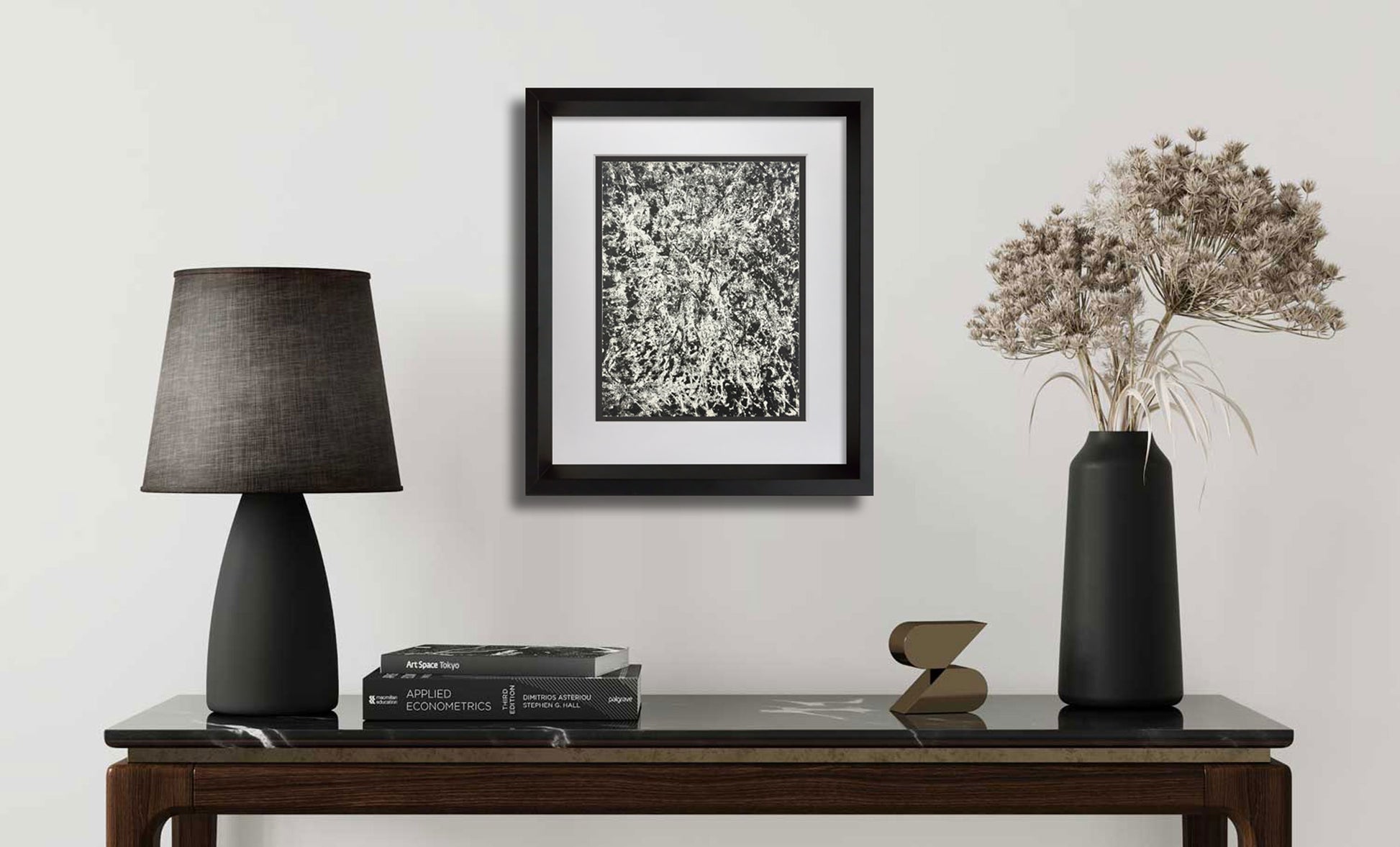 A framed black and white abstract art print hangs on a light gray wall above a dark wood console table styled with a black table lamp, books, a small sculptural object, and a black vase with dried flowers.
