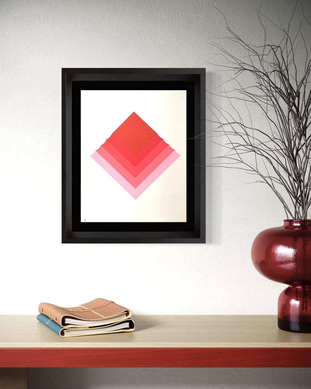 A framed abstract Josef Albers print featuring a geometric diamond pattern in shades of red and pink on a white background, styled on a shelf next to a red vase with dried branches.