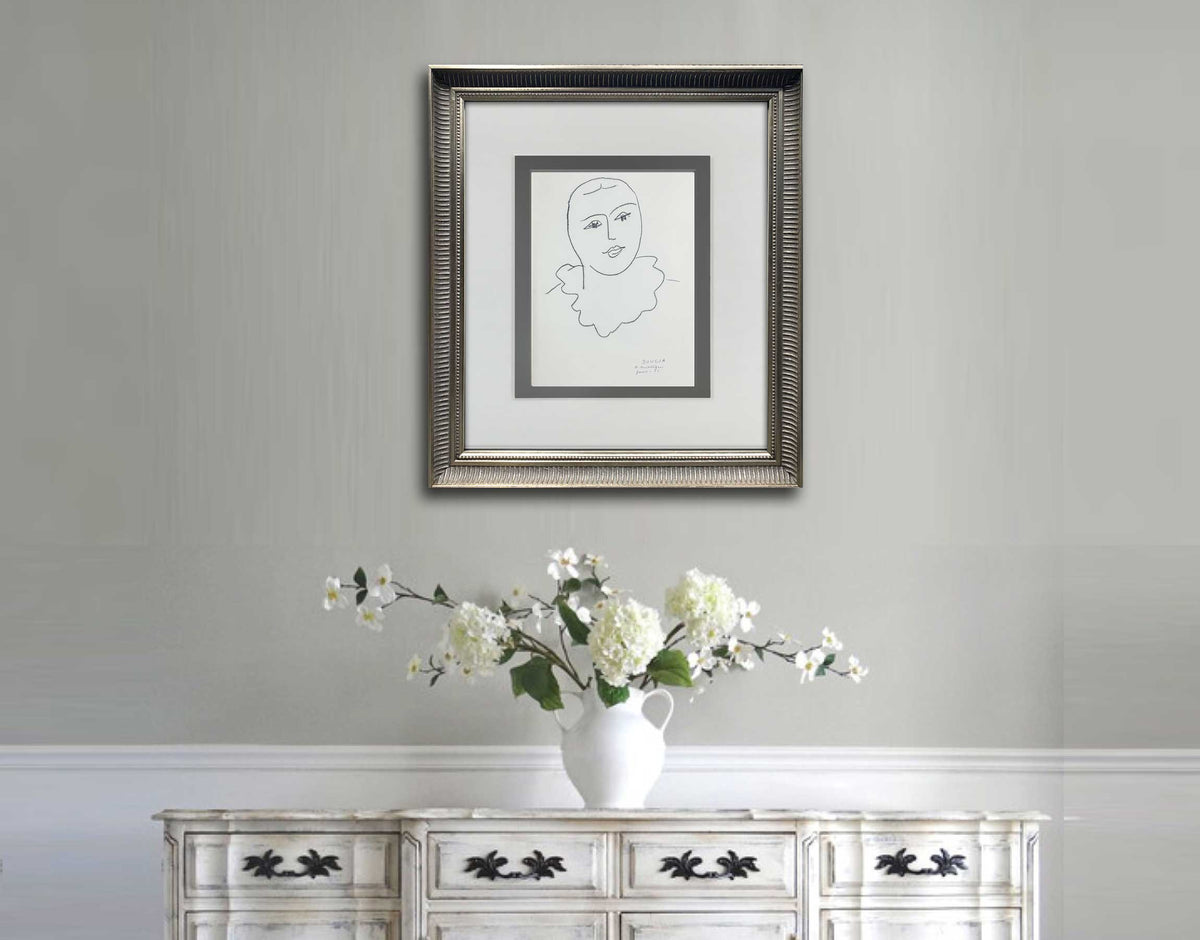 A framed black and white Matisse line art print of a face is displayed on a light gray wall above a white console table with a white vase of flowers.