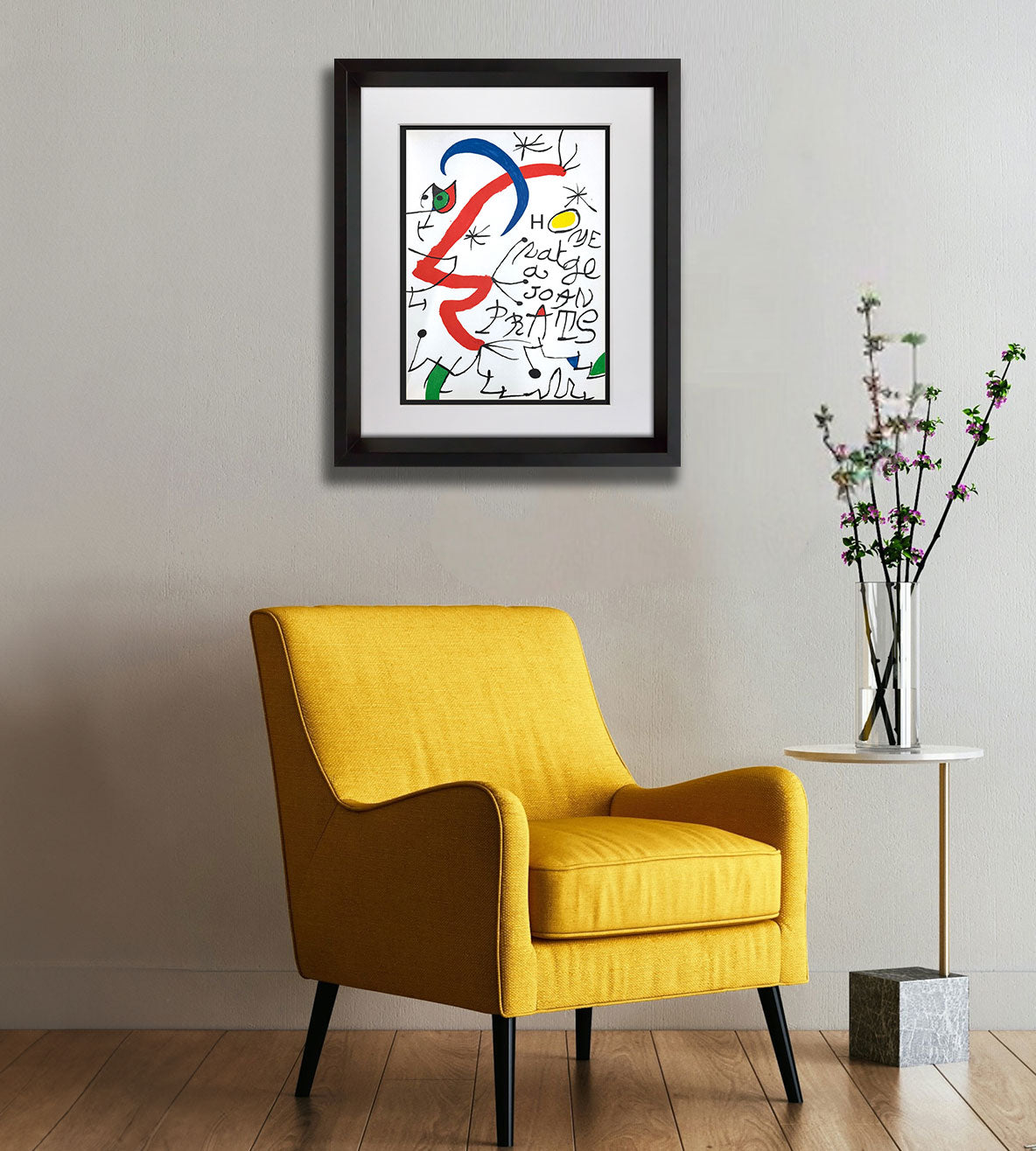 A framed Joan Miro abstract art print with colorful brushstrokes and handwritten text, displayed on a light gray wall above a mustard yellow armchair, with a small side table and vase of branches to the right.