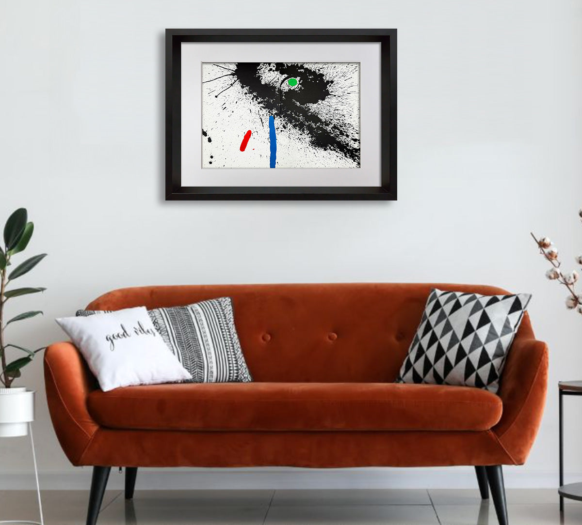 A framed abstract art print with black, white, blue, red, and green brushstrokes hangs on a white wall above a rust-orange velvet sofa with decorative throw pillows.