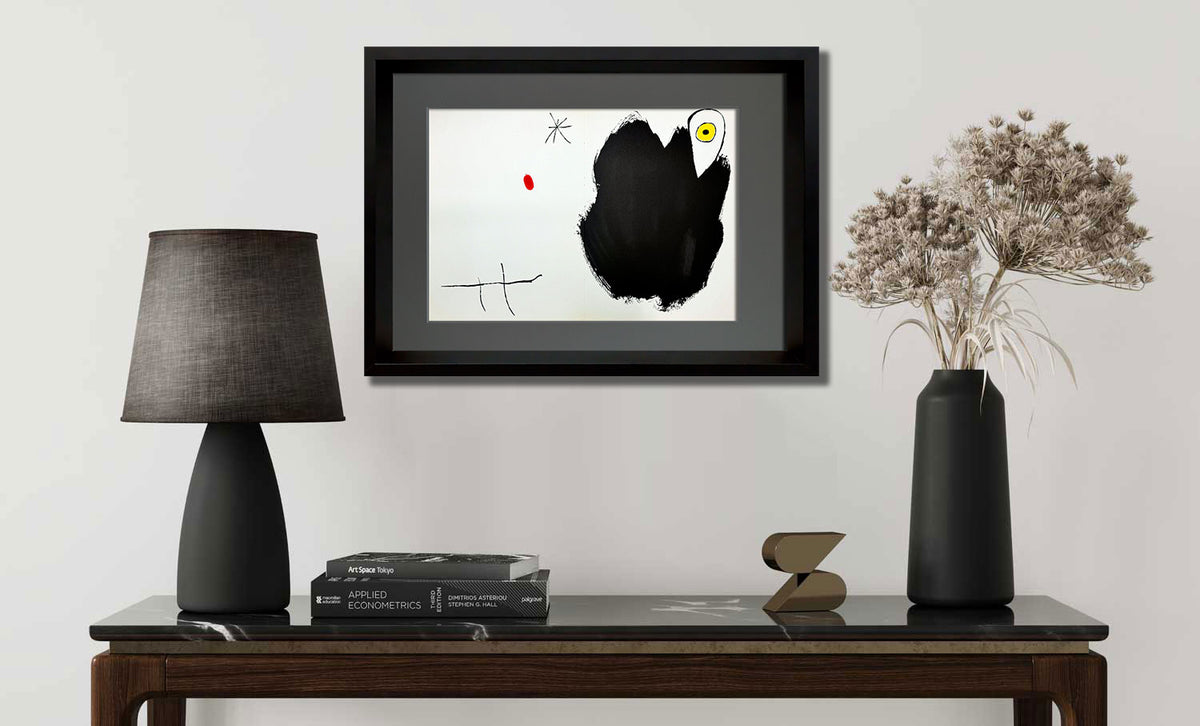A framed abstract art print featuring a large black brushstroke on a white background with small red and yellow accents, displayed on a console table in a styled room setting with a lamp, books, and a vase.