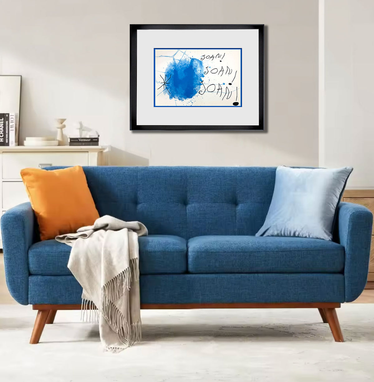 A blue upholstered loveseat with wooden legs in a bright living room, styled with an orange pillow, a light blue pillow, and a beige throw blanket. Above the sofa hangs a black-framed abstract wall art print with a blue paint splatter and handwritten text.