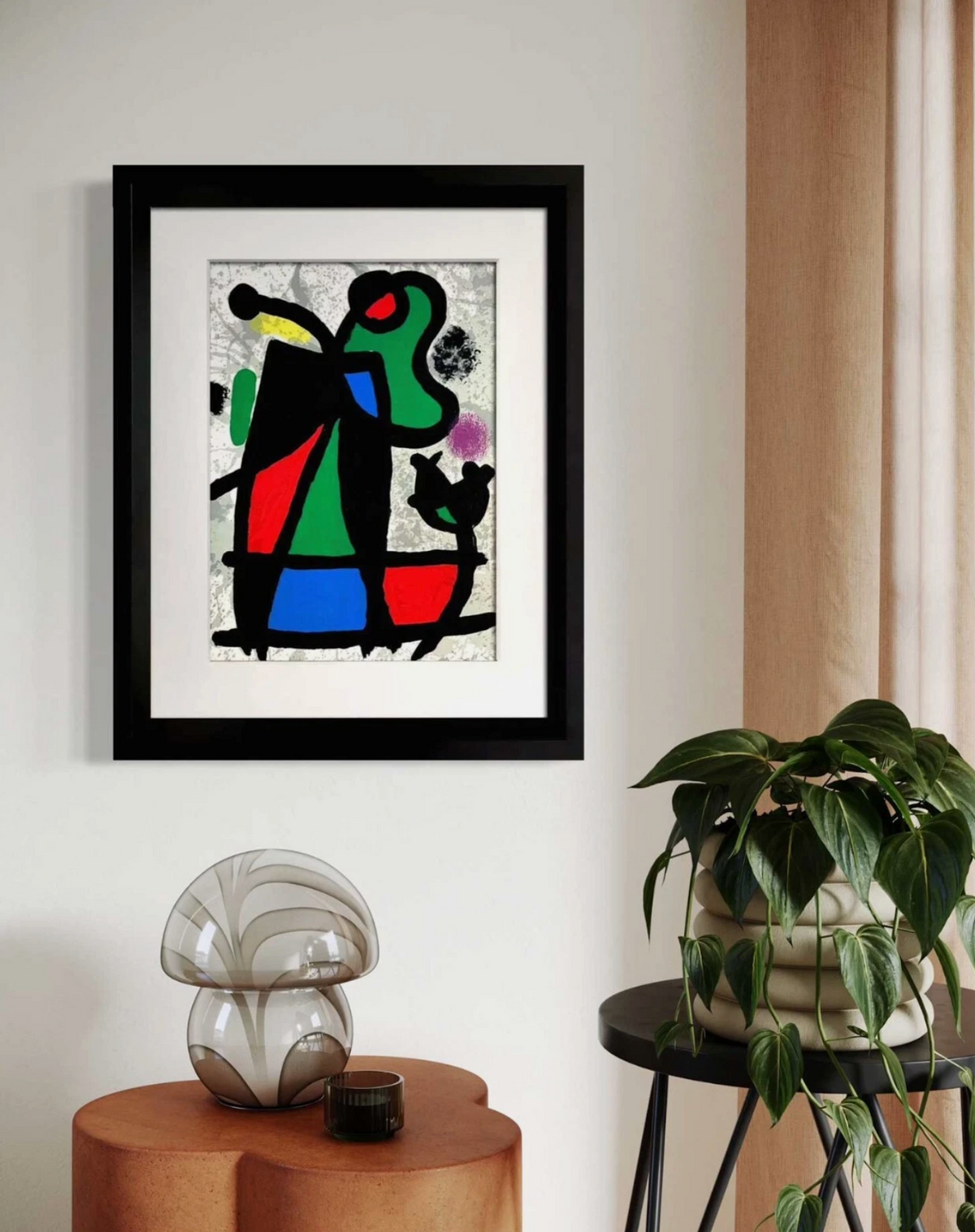 Framed Original Joan Miro abstract wall art hanging on a light wall in a styled interior, with a potted plant and decorative objects below. The artwork features bold black, green, red, blue, and yellow shapes on a textured off-white background.