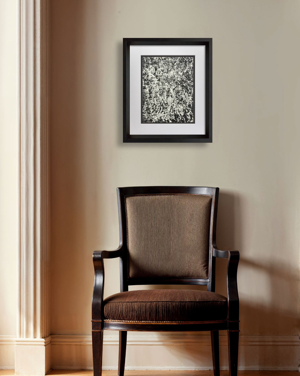 A framed black and white abstract art print with a black frame and white mat, hanging on a beige wall above a dark brown upholstered armchair.