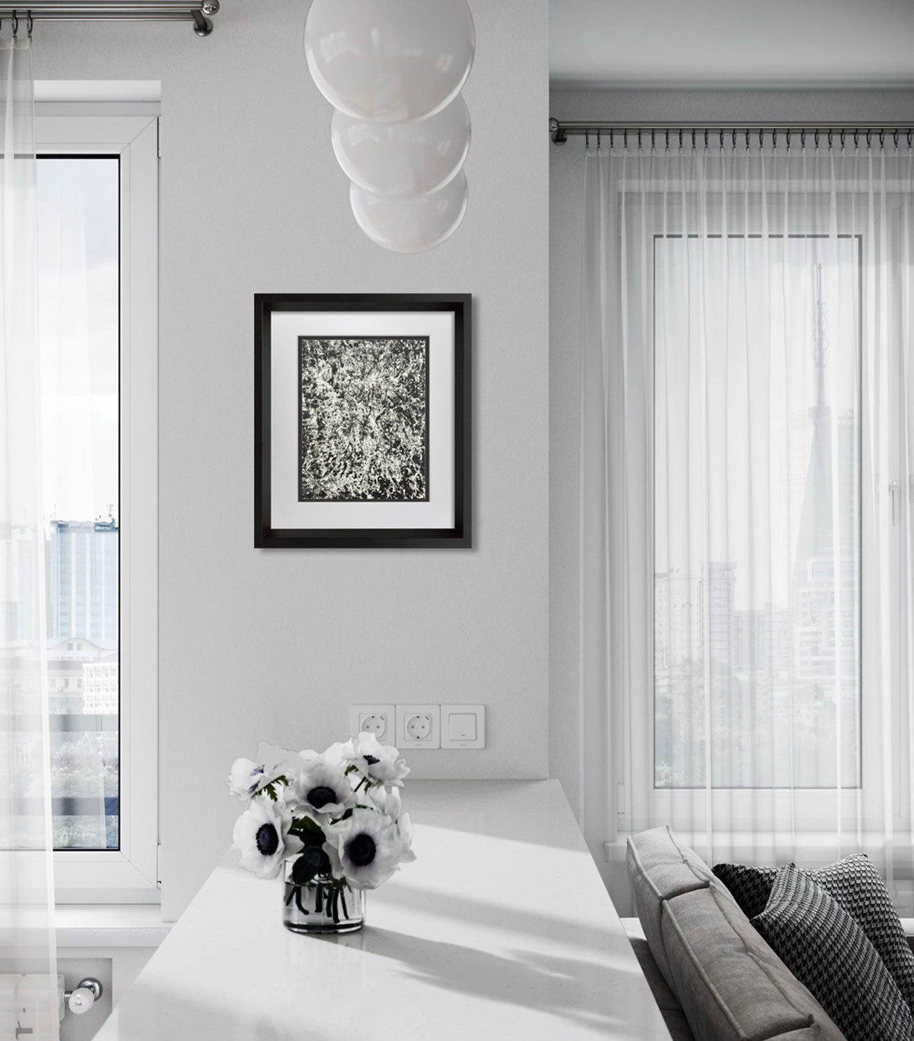 A framed black and white abstract art print hangs on a light gray wall in a modern, sunlit living room with sheer white curtains, a white table with a flower arrangement, and a gray sofa.