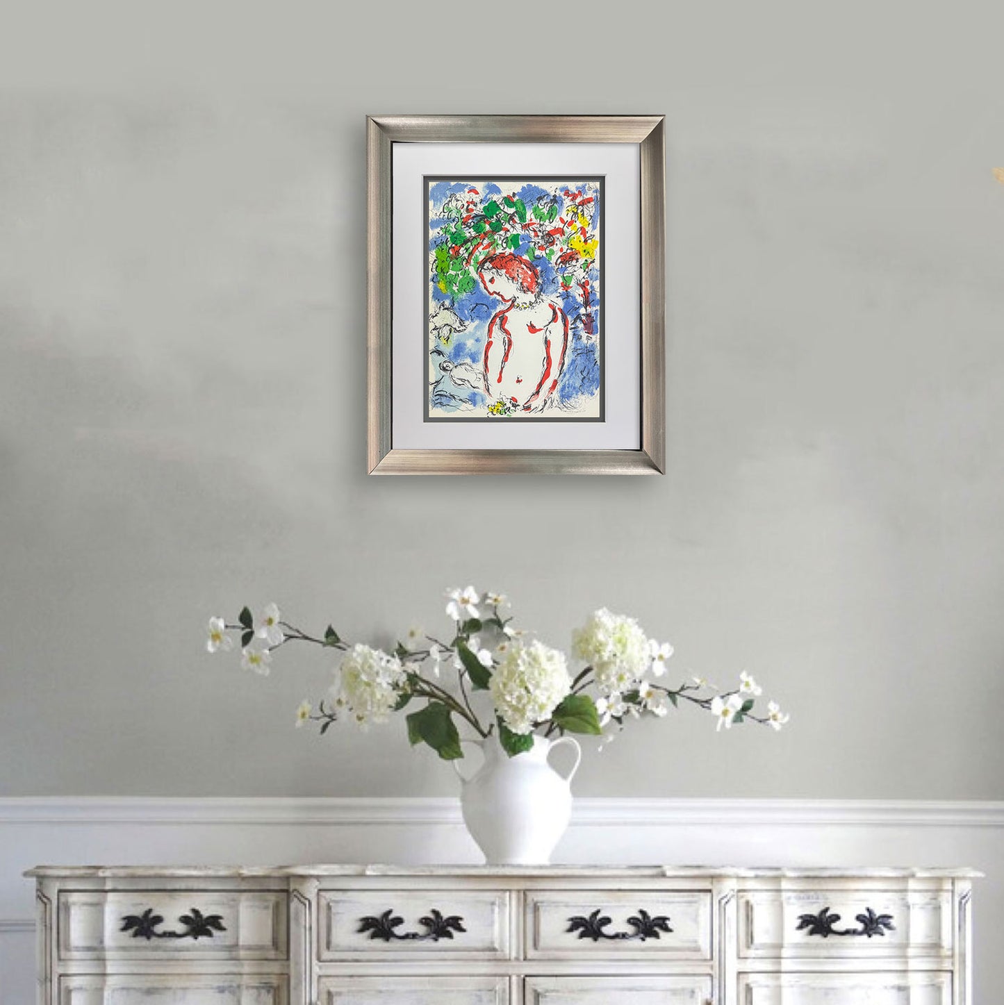 A framed colorful Chagall painting of a woman in a floral landscape, displayed on a light gray wall above a white wooden cabinet with a white vase of flowers.
