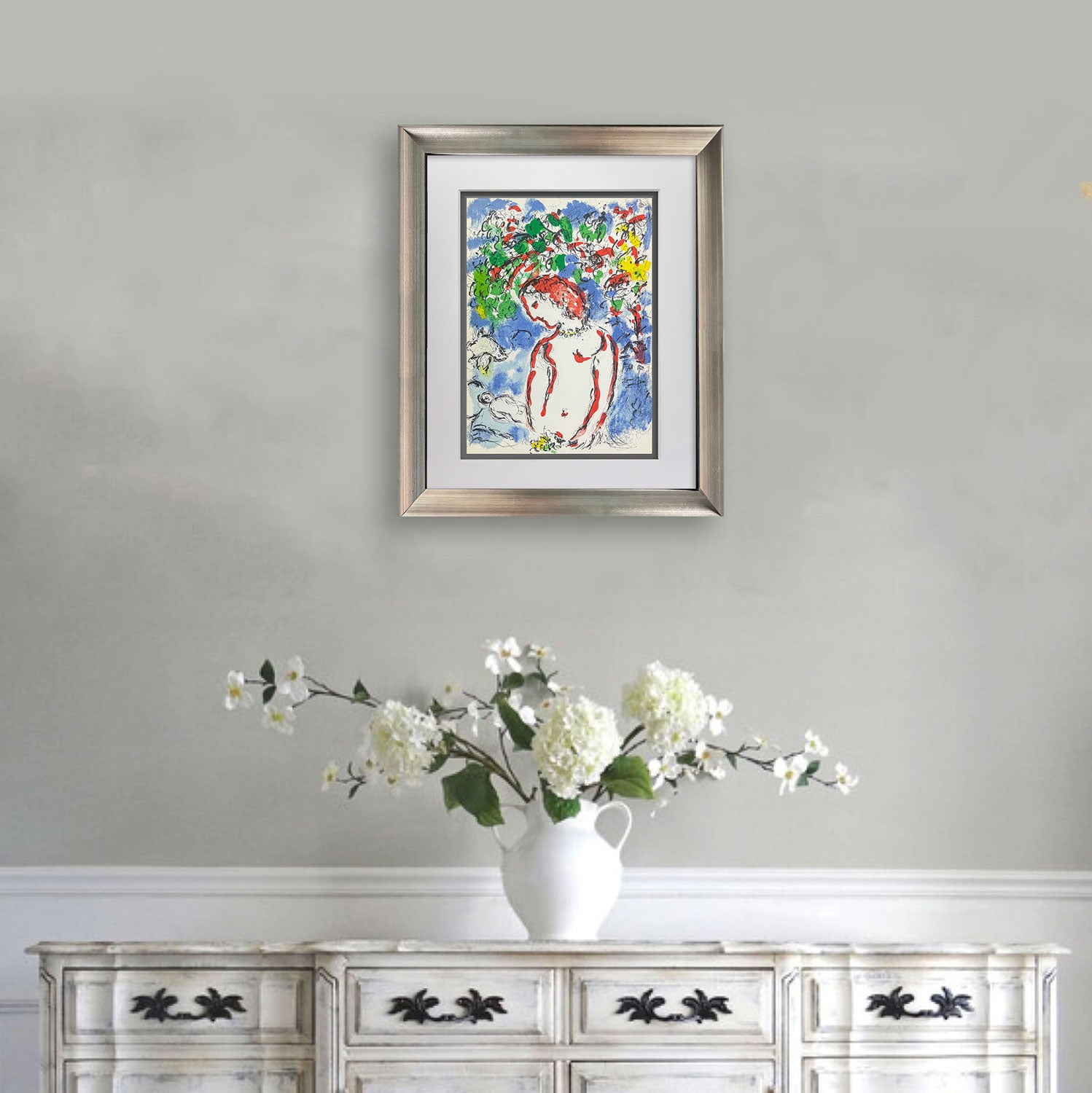 A framed colorful Chagall painting of a woman in a floral landscape, displayed on a light gray wall above a white wooden cabinet with a white vase of flowers.