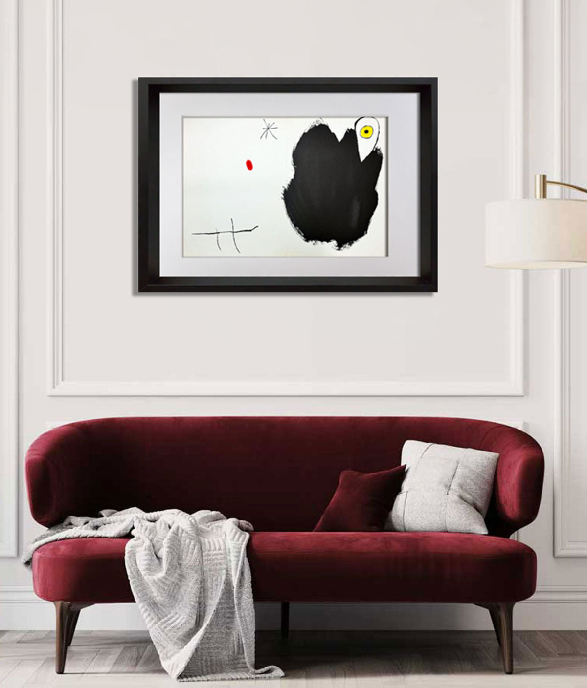 A framed abstract  Joan Miro art print featuring a large black brushstroke on a white background, displayed on a light gray wall above a deep red velvet sofa with a throw blanket.